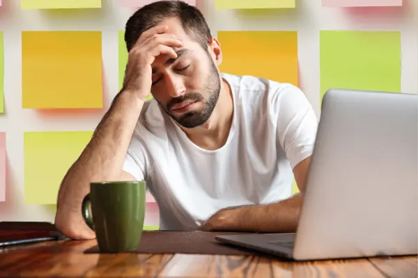 A tired freelancer in a white t-shirt sitting at a desk with a laptop and a green coffee mug, resting his head on his hand, looking stressed. Colorful sticky notes are visible on the wall behind him.