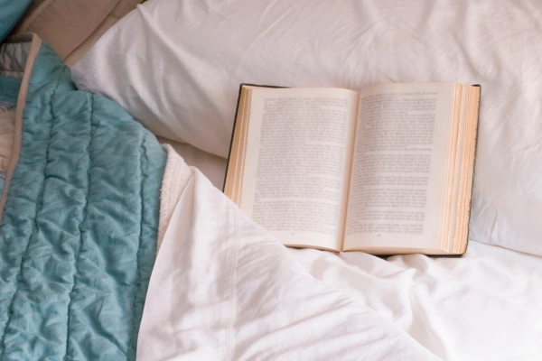 An open book resting on a cozy bed with white sheets and a blue blanket, creating a relaxing bedtime reading atmosphere.