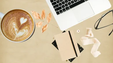 A cozy freelancer workspace with a laptop, a cup of coffee with latte art, a notebook, dried leaves, glasses, and stationery on a warm beige background.