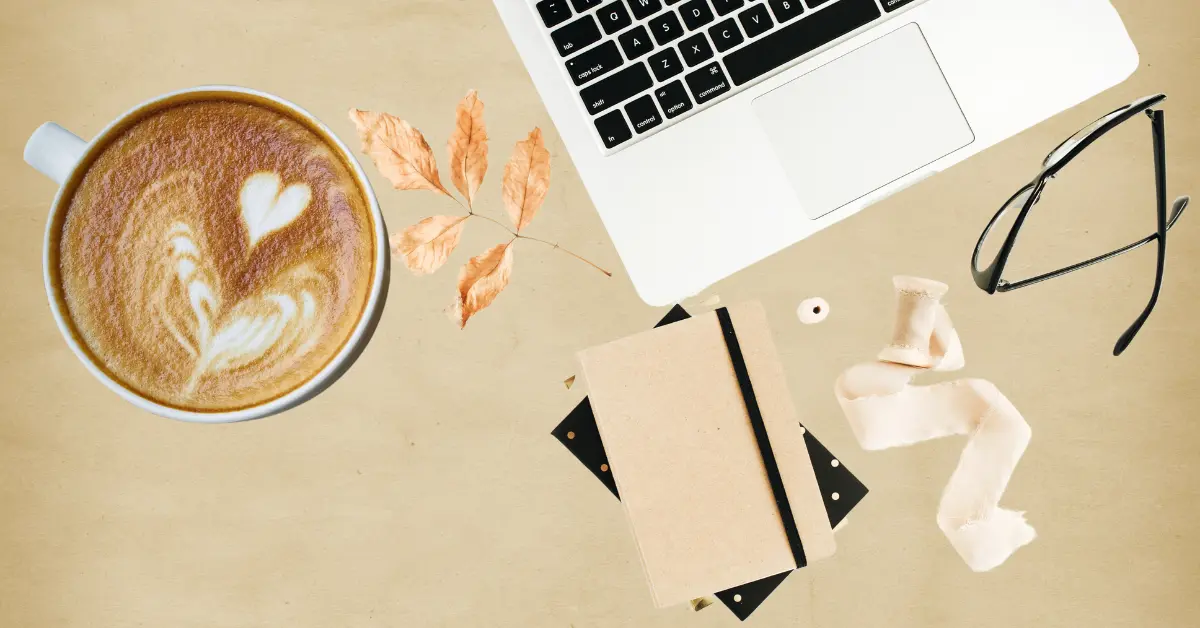 A cozy freelancer workspace with a laptop, a cup of coffee with latte art, a notebook, dried leaves, glasses, and stationery on a warm beige background.